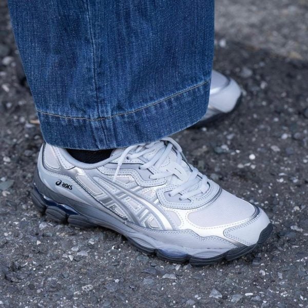 Gray Asics running shoe with white accents on a person wearing blue jeans, standing on pavement.