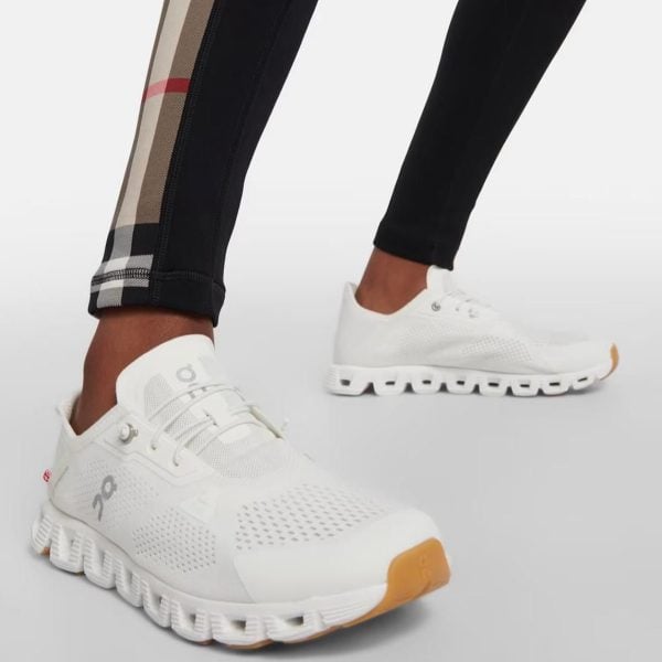 White athletic sneakers with perforated upper and chunky segmented sole, worn by someone in black leggings with a plaid side panel and an orange-tinted outsole at the toe area, shown in a side-view pose.