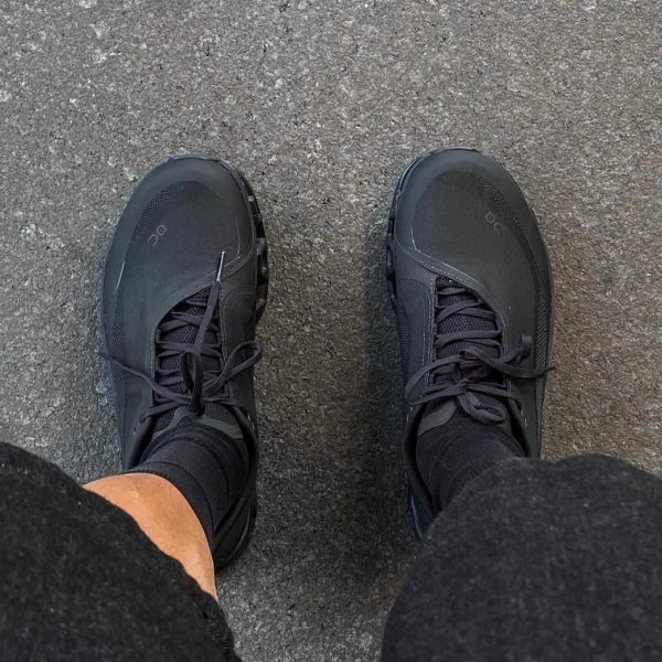 Top-down view of two black athletic sneakers on gray asphalt, seen from above. The laces are tied and the wearer’s legs are in dark pants.