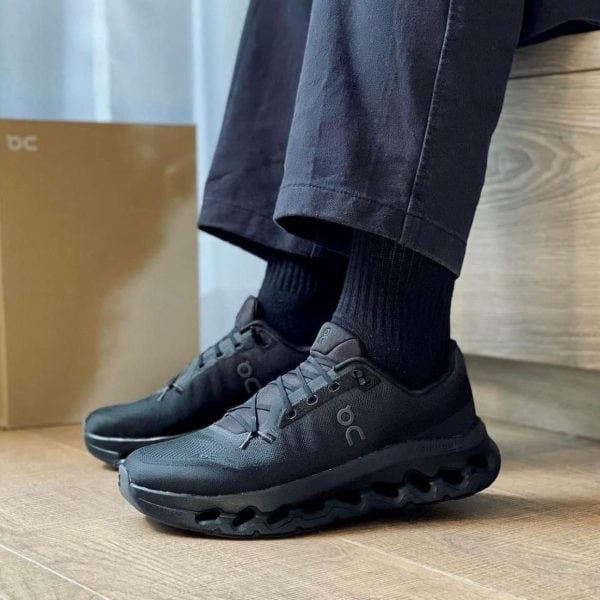Black On running sneakers with a textured upper and distinctive cloud-like sole, worn with black socks and dark pants on a wooden floor indoors.