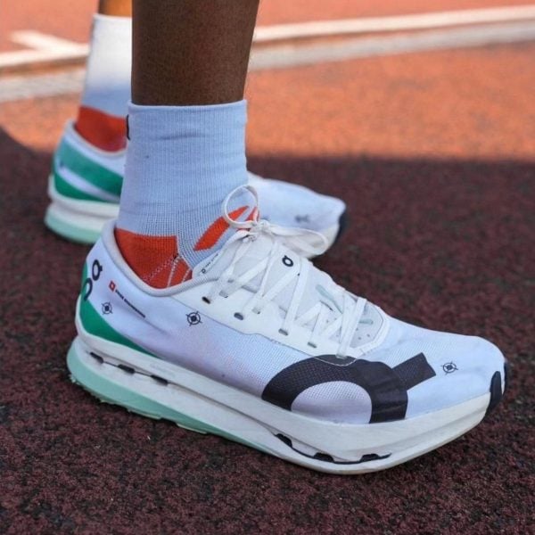 White athletic running shoe with teal, green, and orange accents on a red track, laces tied, paired with light blue socks (close-up).