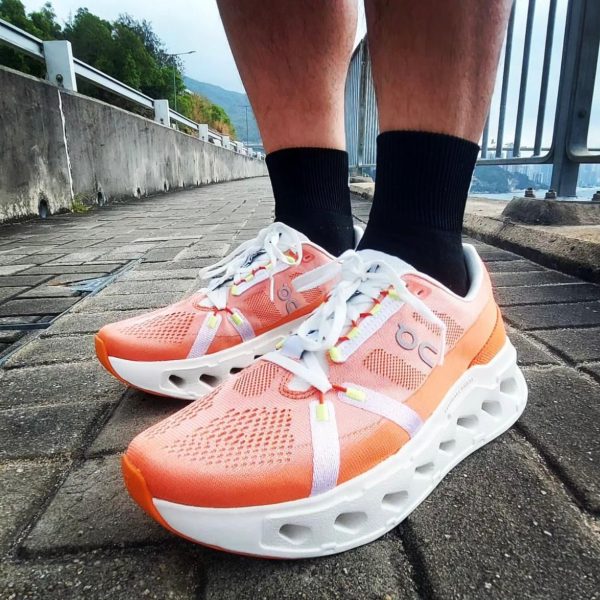 Peach-colored running shoes with white laces and a hollow white sole on a brick walkway beside a railing by the water.
