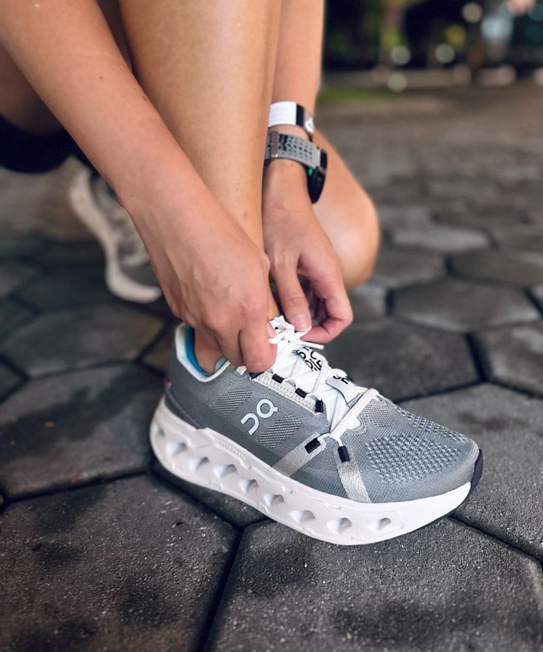 WhatsApp_Image_2025-01-16_at_15.08.53 | AE JERSEY Person ties white laces on a gray On running sneaker while kneeling on a cobblestone pavement.