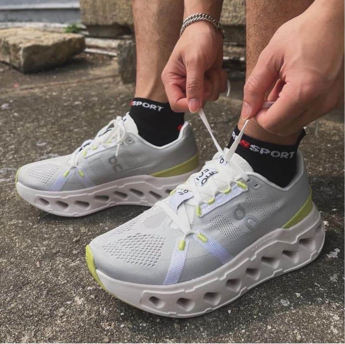 WhatsApp_Image_2025-01-18_at_13.18.20 | AE JERSEY Person tying the laces of white running shoes with neon yellow accents on a concrete surface.