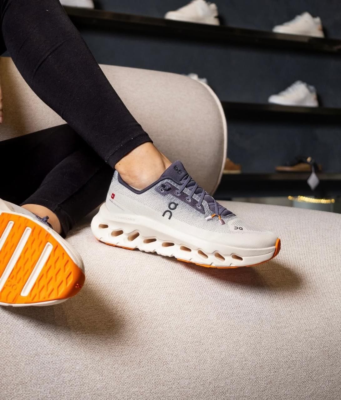 WhatsApp_Image_2025-01-22_at_13.03.22 | AE JERSEY Close-up of a gray On running sneaker with an orange sole resting on a beige sofa, worn by a person seated indoors.