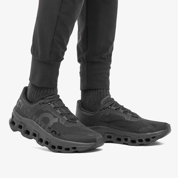 Black athletic sneakers with a vented, cube-cut sole and knit upper, shown on a person wearing black pants and socks.