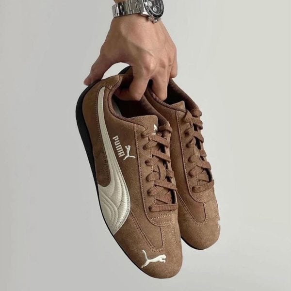 WhatsApp_Image_2025-01-28_at_10.04.04 | AE JERSEY Brown suede Puma sneakers with white logo held by a hand wearing a silver watch, against a neutral background.