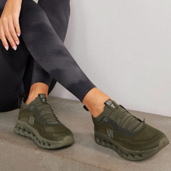 Person seated on a concrete step wearing olive-green athletic sneakers with black patterned leggings. The shoes have a unique chunky sole and laces visible.