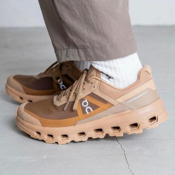 Tan/orange On running shoes with a chunky sole, white socks, and light brown pants on a gray floor.