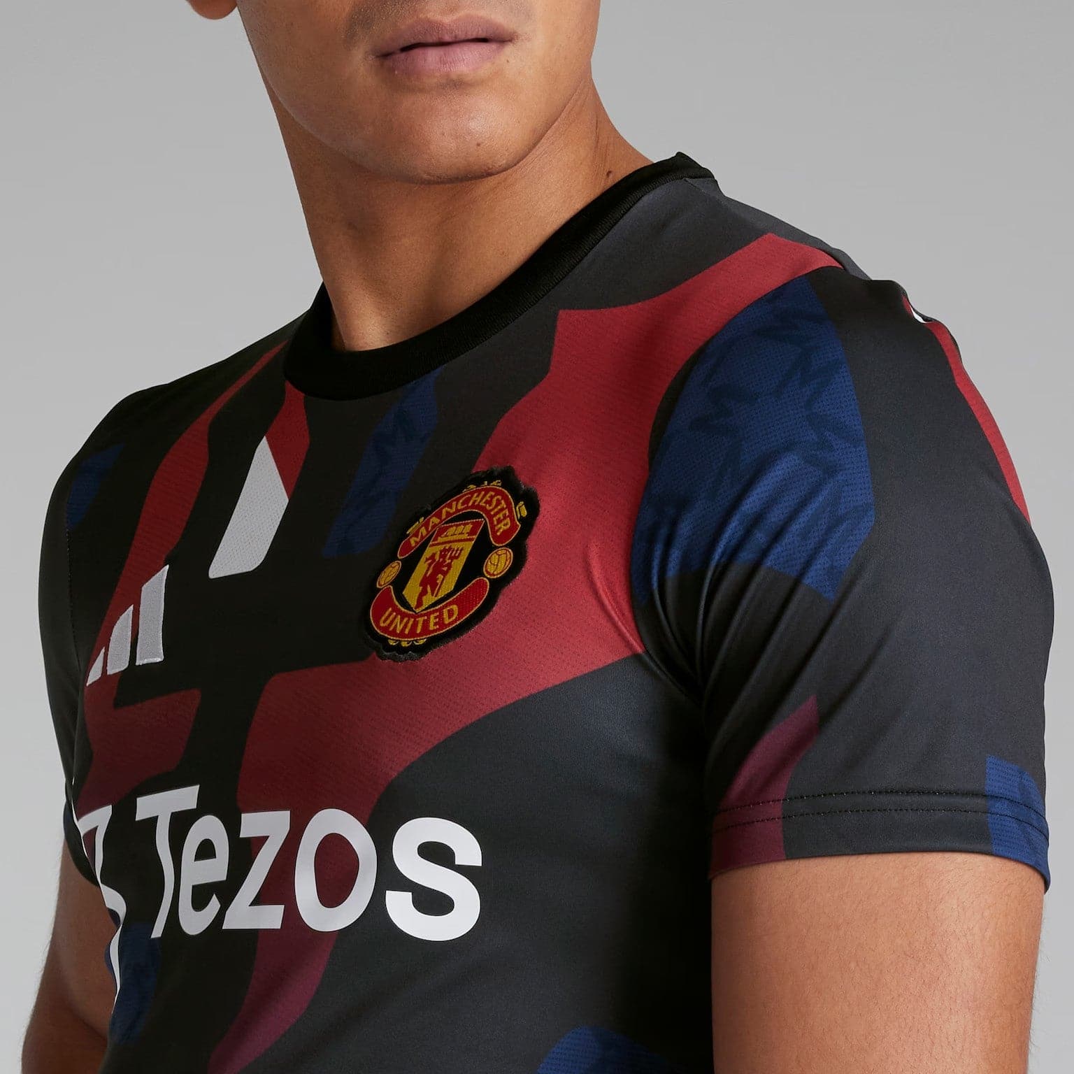 YFlKtRonDhEhvyS | AE JERSEY Close-up of a Manchester United football jersey with red and black diagonal stripes, showing the club crest and the Tezos sponsor logo on the chest area.