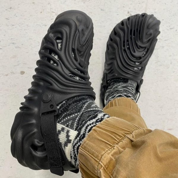 Black chunky sandals with cut-out, wave-like sole viewed from above, worn with patterned socks and tan pants.