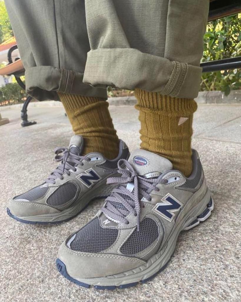 a61f1e97-fbc5-4a4b-9652-4a09227c3f55 | AE JERSEY Close-up of gray New Balance sneakers with navy N logos, mustard socks, and rolled khaki pants outdoors on a concrete surface.