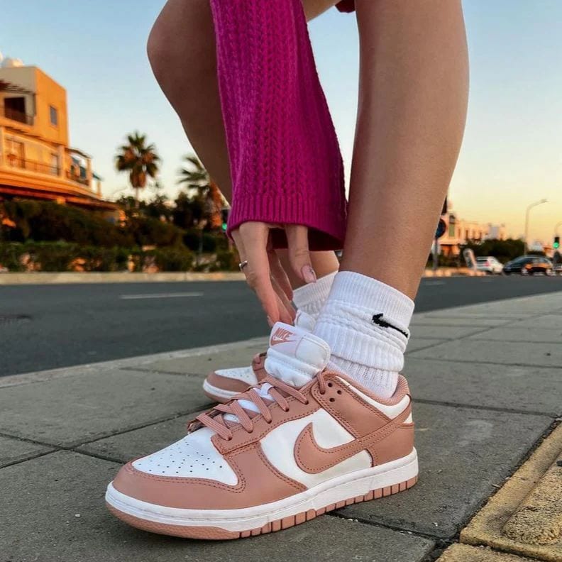 a62fdabf-28ae-4393-9b80-a40313956751 | AE JERSEY Close-up of white and pink Nike sneakers on a person’s feet, standing on a sidewalk with a city street in the background at sunset.