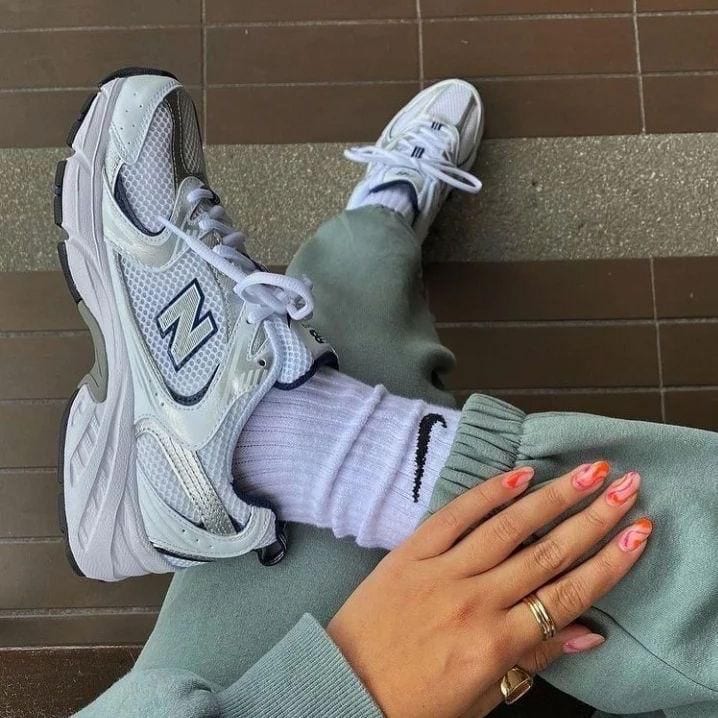a72287ee-425c-49b9-9ce2-fa50a400c69a_ae43de14-9ce2-40d0-8725-fed6ac6a2319 | AE JERSEY White New Balance sneakers with thick soles, white socks, sage-green joggers, and a hand with pink nail polish resting on the knee on a brown tile floor.
