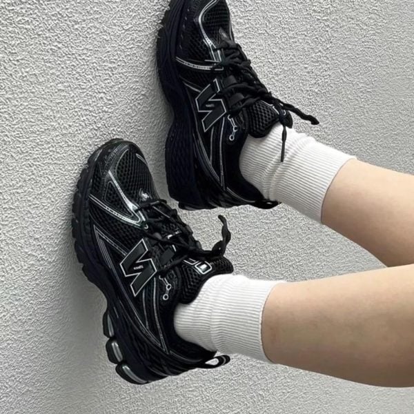 Two black athletic sneakers with chunky soles and white detailing rest against a textured gray wall, worn with white socks.