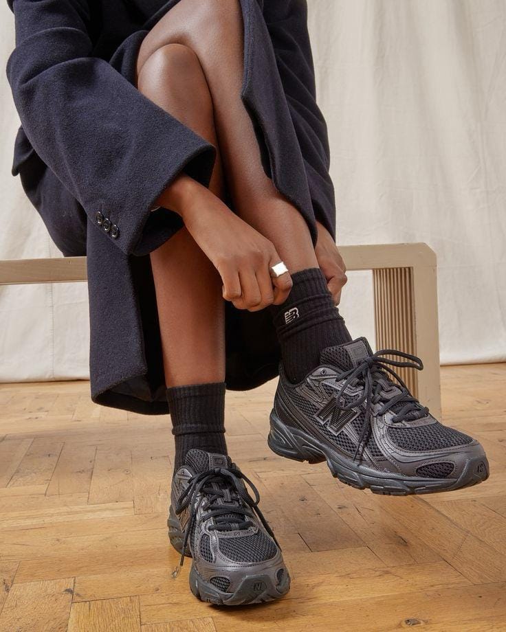 aa197772-67be-417a-8dea-ca96a996c010 | AE JERSEY Person in a dark coat sits on a wooden floor and ties black athletic sneakers with chunky soles and black socks.