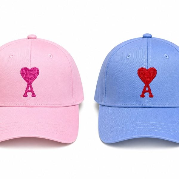 Two baseball caps side by side: pink on the left and blue on the right, each with a red heart-shaped ribbon embroidery on the front of the crown.