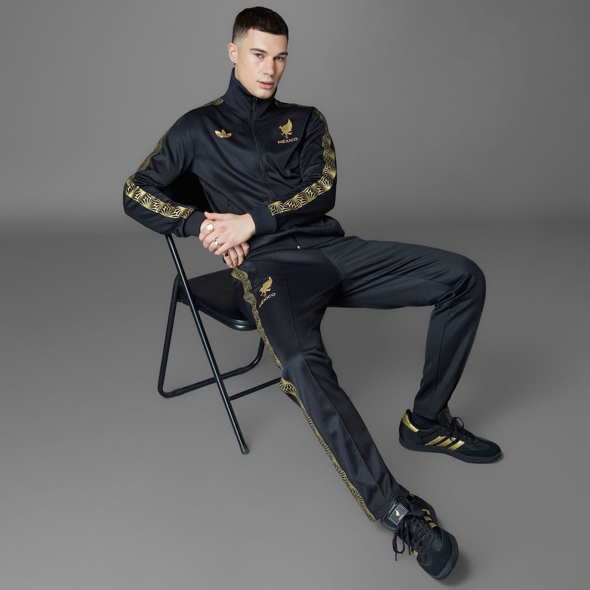 adidas-Samba-Mexico-Black-Gold-JH5656 | AE JERSEY Man in a black tracksuit with gold detailing sitting on a folding chair, facing the camera.