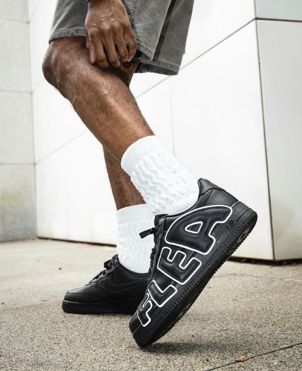 b018f503-2d0f-4ba6-99b9-1293841a140b | AE JERSEY Person wearing black FILA sneakers with white detailing and large 'FILA' lettering, walking outdoors in white socks and gray shorts.