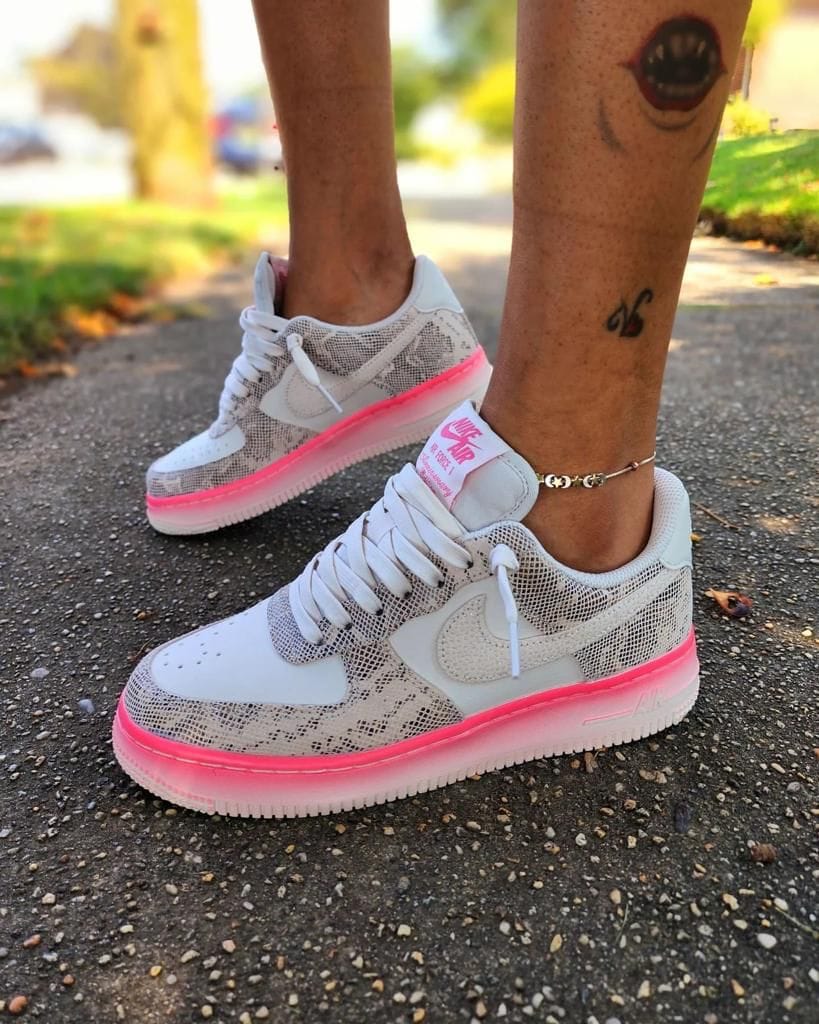 b0d424a4-e7ba-4729-a6c3-0283bbb9b7aa_d3ce534e-1b44-4603-864b-0ae88ba264db | AE JERSEY White sneakers with a bright pink sole on a person standing on a paved sidewalk; visible ankle tattoo and bracelet.