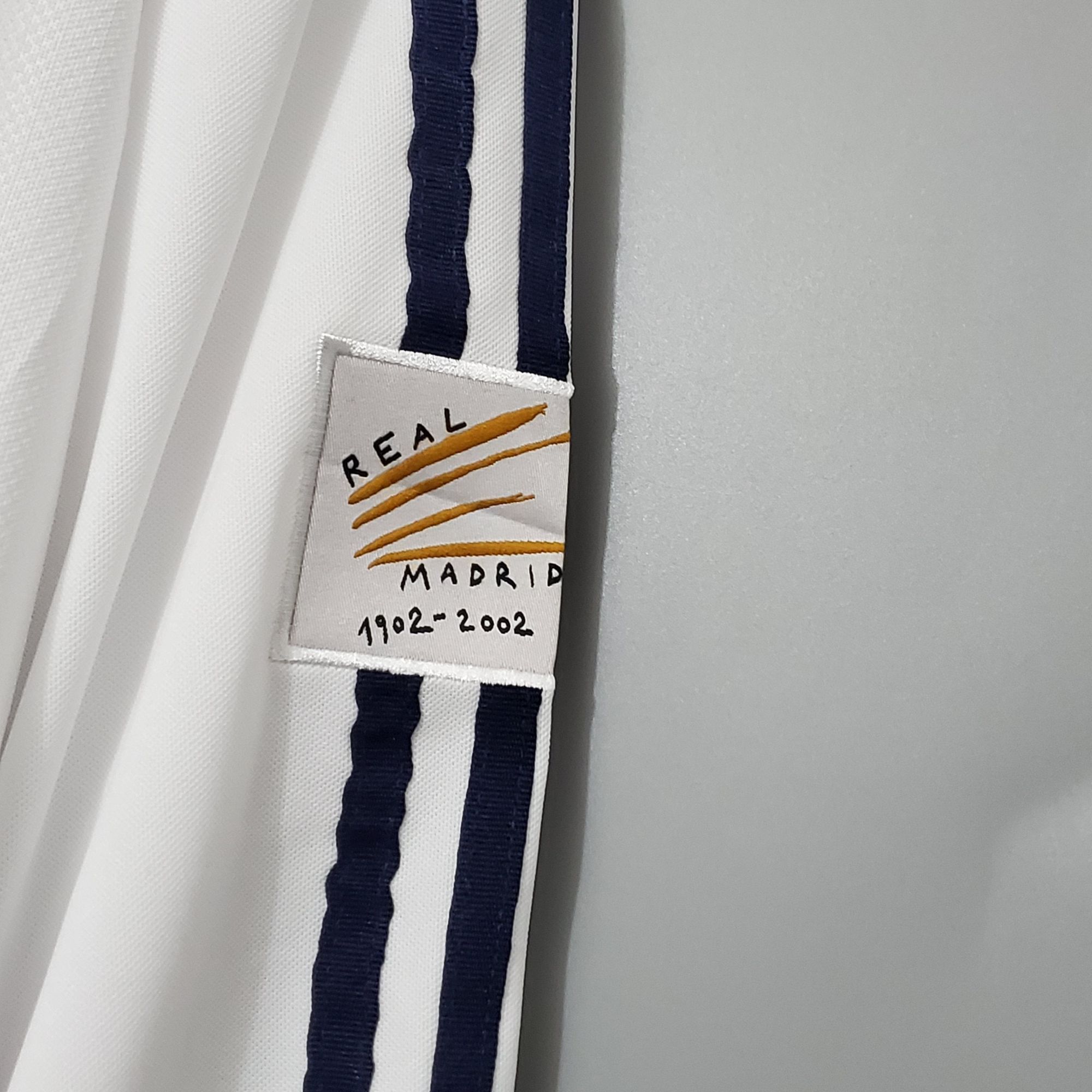 b1f3d17a_fc8eb008-93bc-401b-a9df-a04b7afa3a2d | AE JERSEY White fabric with a dark blue stripe and a small tag reading 'REAL MADRID 1902-2002', suggesting a commemorative scarf or ribbon.