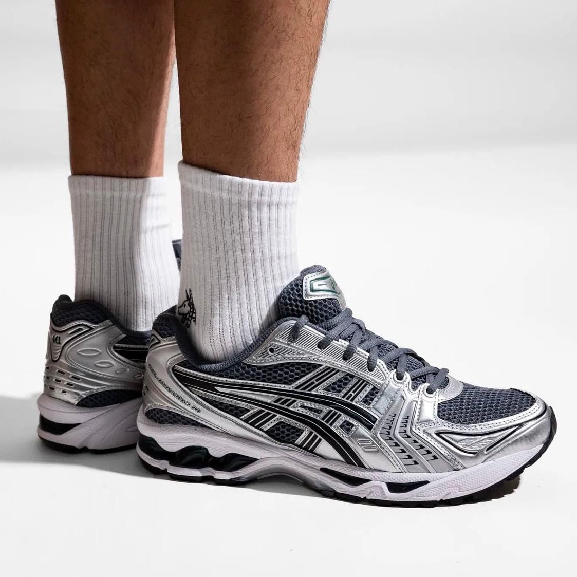 b3195f73-c064-41de-942c-e645bc5bf041 | AE JERSEY Close-up of a person wearing white crew socks and silver/black running shoes standing on a white surface, showing the sneakers' details.