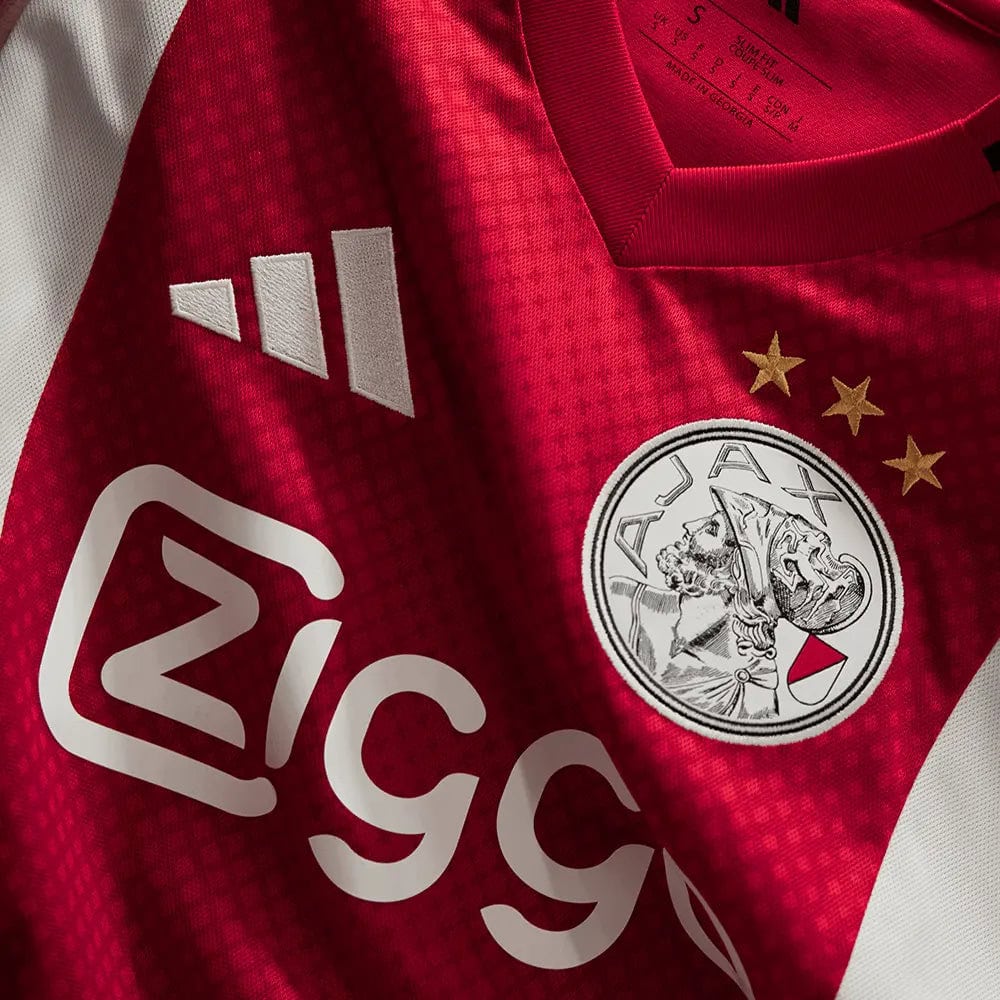 b3e0392f-f64d-46cb-507f-9673ac1a718d | AE JERSEY Close-up of a red soccer jersey featuring a white sponsor logo and a circular crest with a knight and three gold stars.