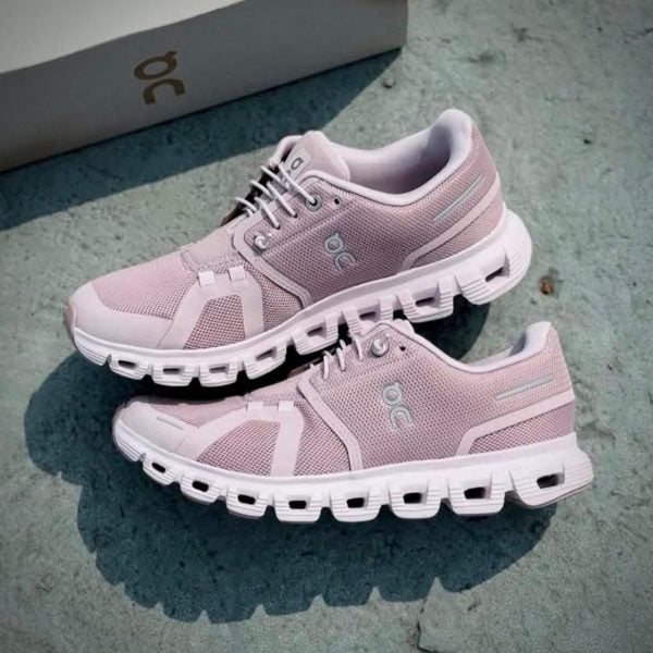 Light pink running shoes with chunky white soles resting on a concrete surface, a box in the background; brand logo visible on the side.