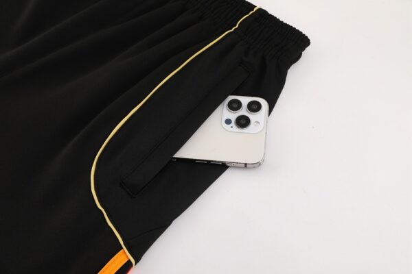 b81620de-600x400-1 | AE JERSEY Black athletic shorts with gold piping; a white smartphone peeks out of the front pocket on a light surface.