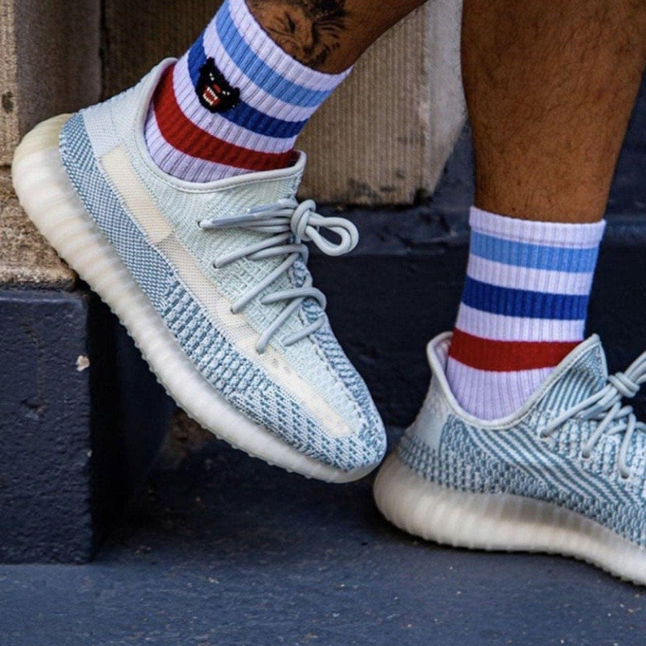 b9c522e9-d856-4295-ba95-dc0685165cf9_30d70353-cdff-4eb4-8c42-338b6cf94b18 | AE JERSEY Light blue knit sneakers with pale soles, laced, worn with red, white, and blue striped crew socks on a dark step.