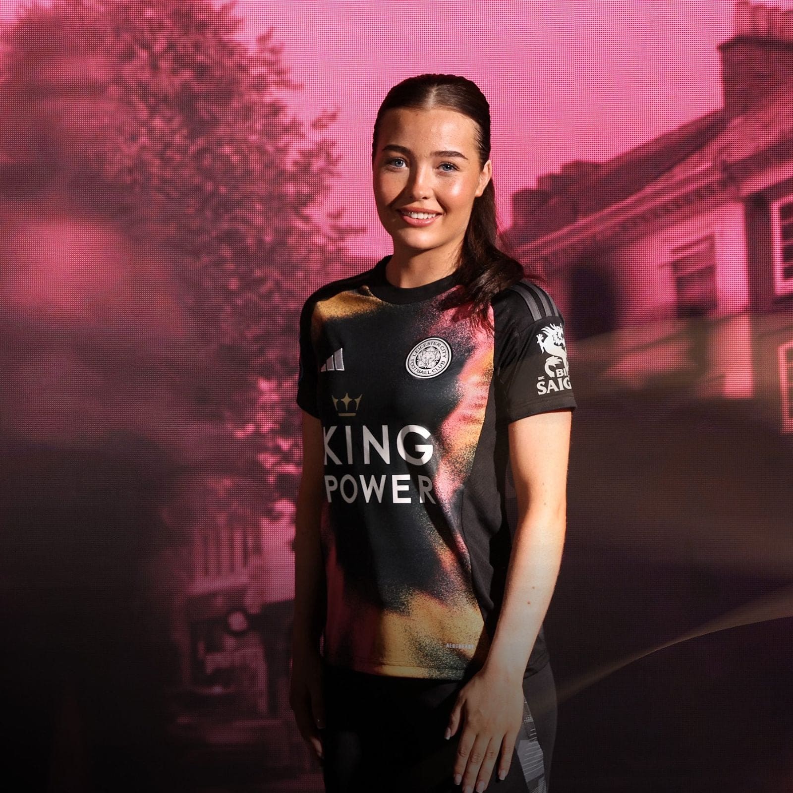 bDl42CKfAzRZQzW | AE JERSEY Smiling woman in a black Leicester City football jersey with a King Power logo, standing against a pink backdrop.