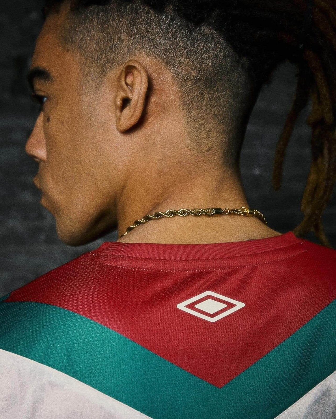 bYeiHOHRFtDusXW | AE JERSEY Profile of a person with a short fade and dreadlocks, wearing a red and teal geometric sports jersey and a gold chain necklace