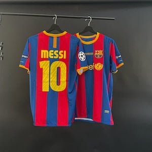 barcelona-classic-201011-final-with-messi-10-badges-jersey-443563 | AE JERSEY Two FC Barcelona jerseys hanging on hangers, blue and red vertical stripes; one shows 'MESSI 10' on the back, the other features the Barca crest and patches.