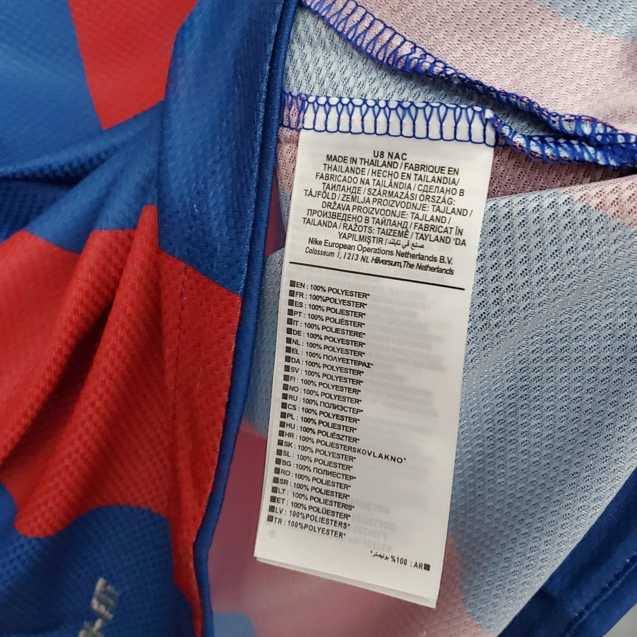 barcelona-classic-201011-final-with-messi-10-badges-jersey-877067 | AE JERSEY Close-up of a white clothing tag on a blue garment, showing origin and fiber details (Made in Thailand, polyester) with blue overlock stitching nearby.