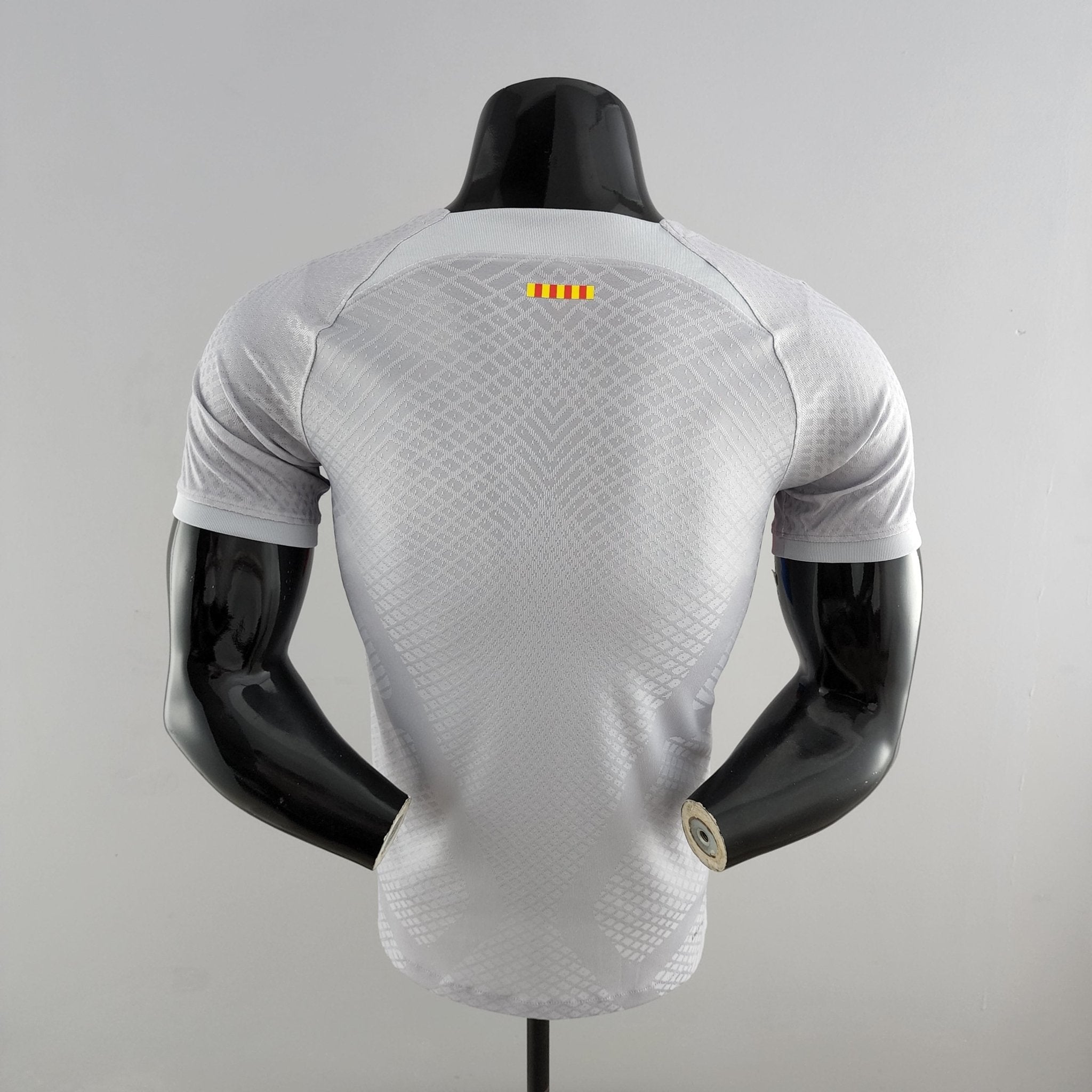 barcelona-third-player-version-jersey-202223-700272 | AE JERSEY Back view of a gray textured athletic shirt on a black display mannequin, with a small yellow and red striped label on the upper back.