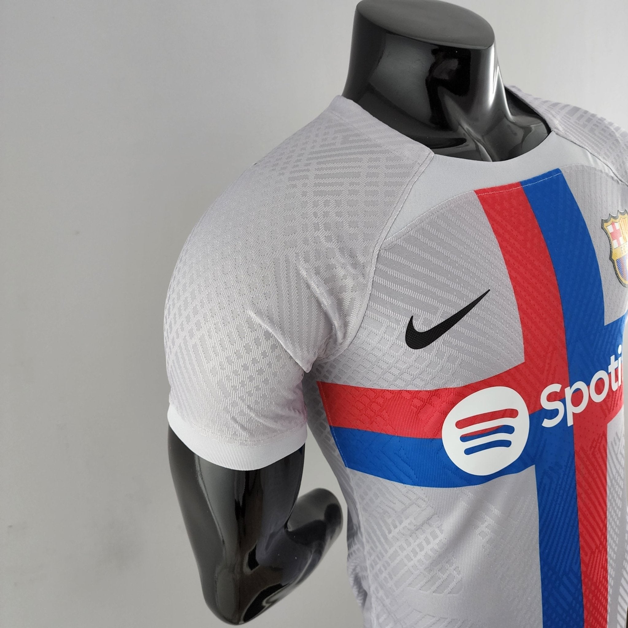barcelona-third-player-version-jersey-202223-923789 | AE JERSEY Mannequin wearing a light gray sports jersey with red and blue vertical stripes and a crest on the chest; only upper body visible.