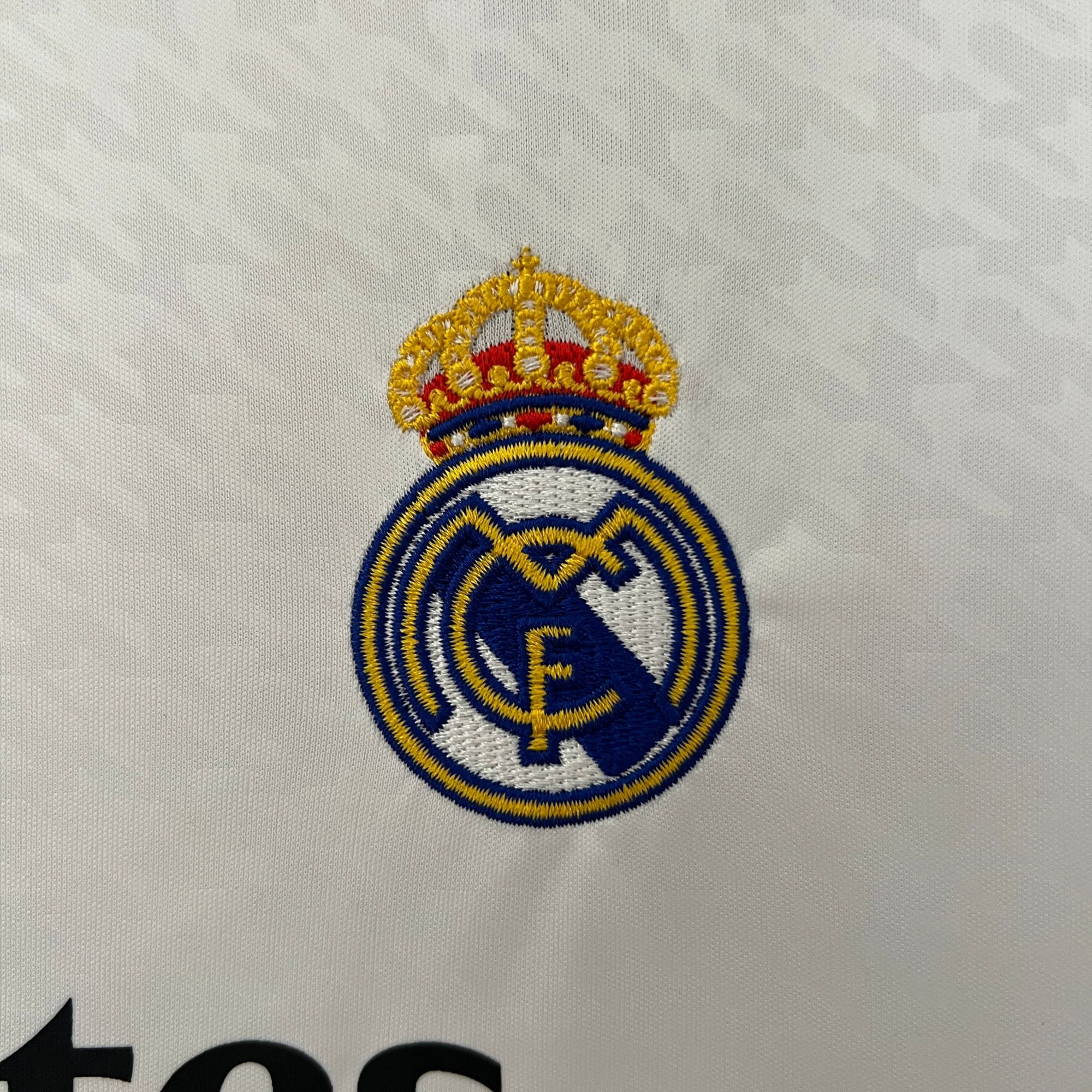bec6f897 | AE JERSEY Partial embroidered crest featuring a gold crown above blue and white elements on white fabric, top portion visible