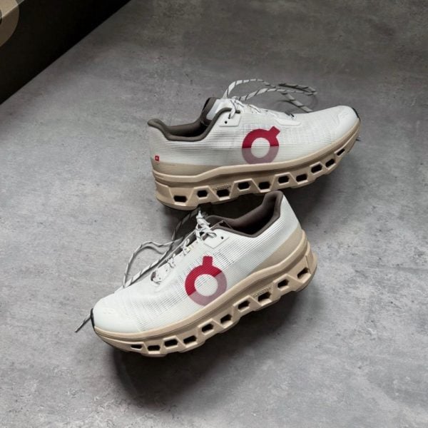 Pair of white and beige running shoes with a chunky, hollowed-out beige sole and red circular logo on the side, lying on a gray floor; laces loose.