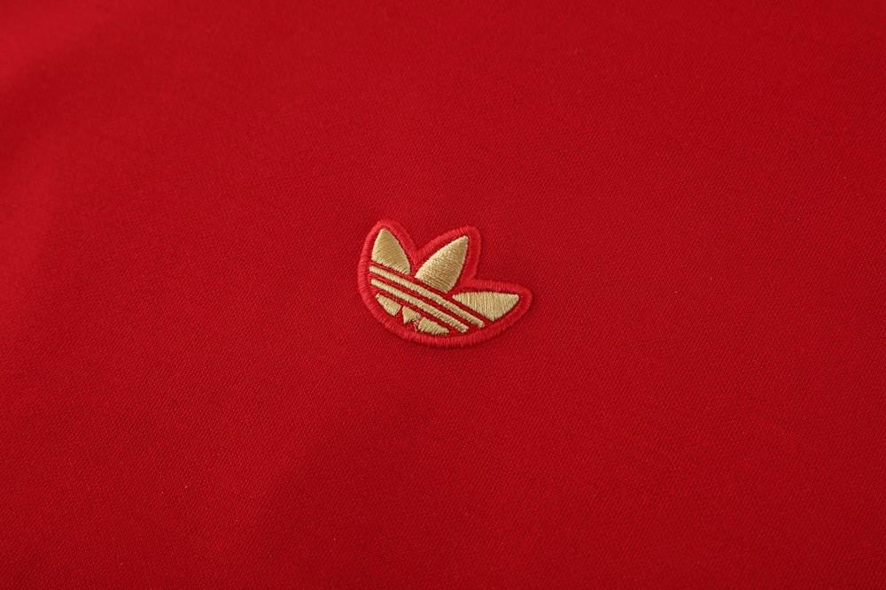 c273ed41-e2e7-43af-b6f6-2eab38a0887d | AE JERSEY Close-up of red fabric with a small gold Adidas trefoil logo embroidery.