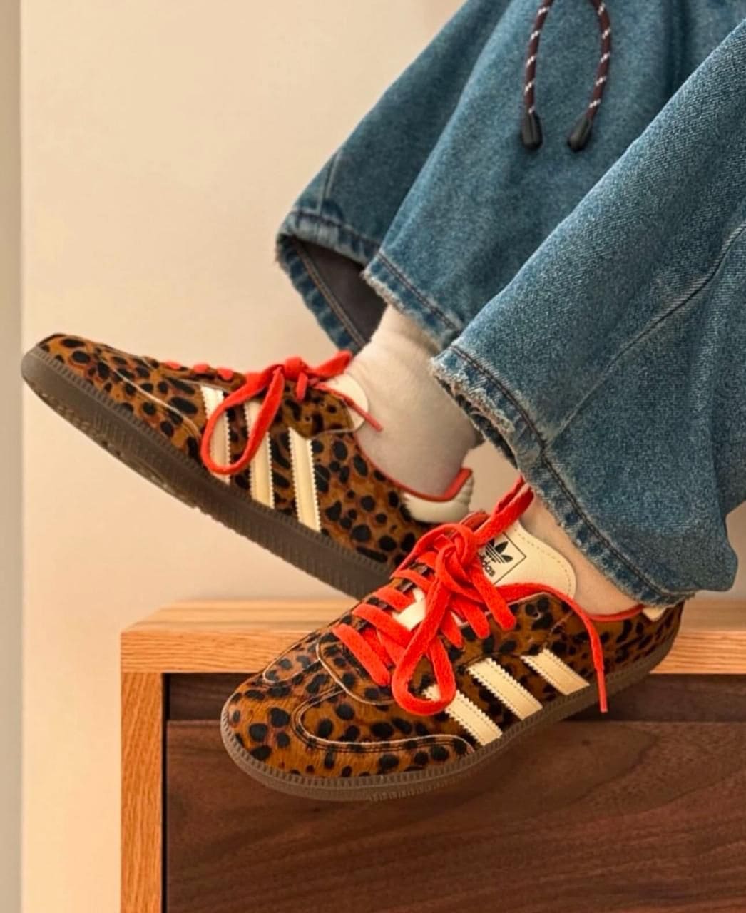 c4507a2c-0f73-4d0f-9855-08b21c6014ab | AE JERSEY Leopard print sneakers with red laces and white stripes resting on a wooden surface, worn with jeans and white socks.