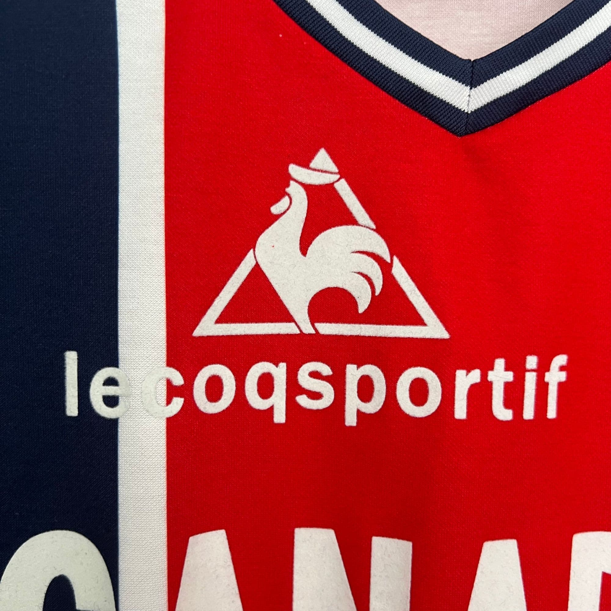 c45d1cee_569ad016-3ce8-4b7a-9494-9d749fa226d1 | AE JERSEY Close-up of red fabric showing part of the Le Coq Sportif logo — a white triangle with a white rooster silhouette and partial brand text visible on the red background.