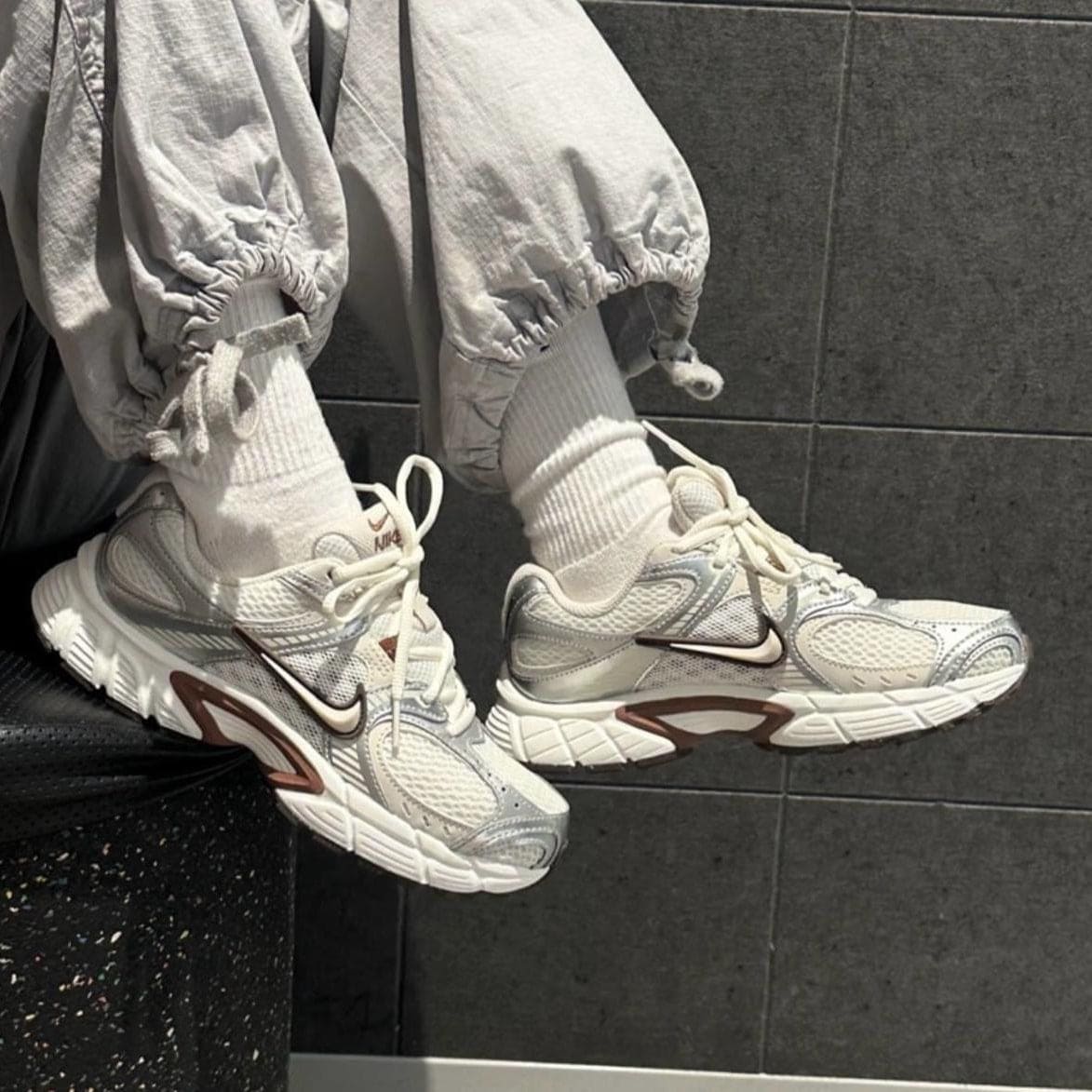 c4aa9707-9f8c-4855-9dbf-3451e7cd198b | AE JERSEY Close-up of white Nike sneakers on feet wearing white socks and light gray joggers, against a dark tiled wall.