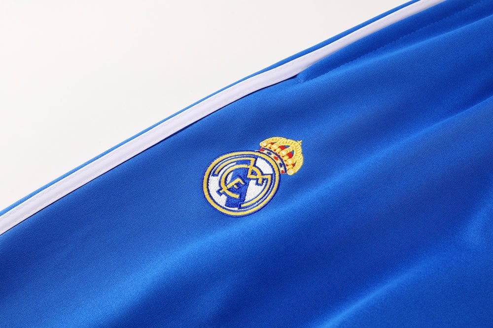 c909c46d | AE JERSEY Close-up of blue satin fabric featuring the Real Madrid club crest embroidered in gold and white with a white stripe along the edge.