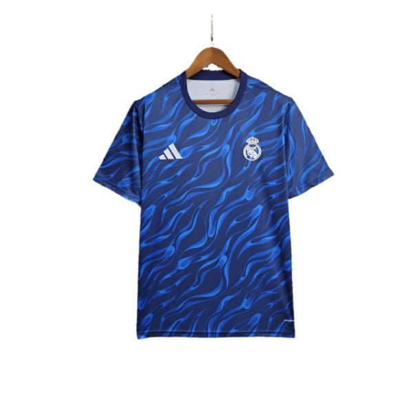 Blue Real Madrid football jersey with a wavy pattern, crest on the left chest and Adidas logo on the right, hanging on a wooden hanger.