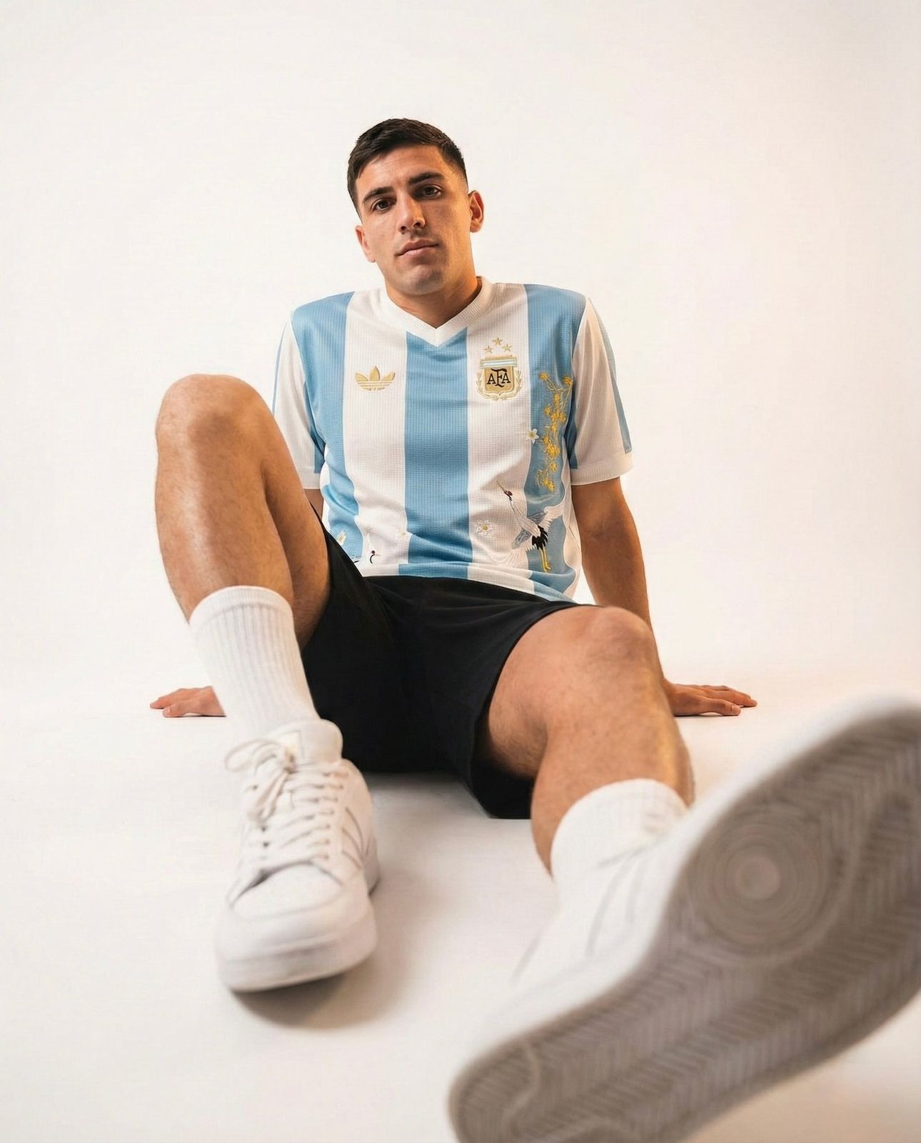 d011a91e-ea11-4685-9800-954252c27236 | AE JERSEY Young man wearing an Argentina football jersey sits on the floor with one leg extended toward the camera against a plain white background.