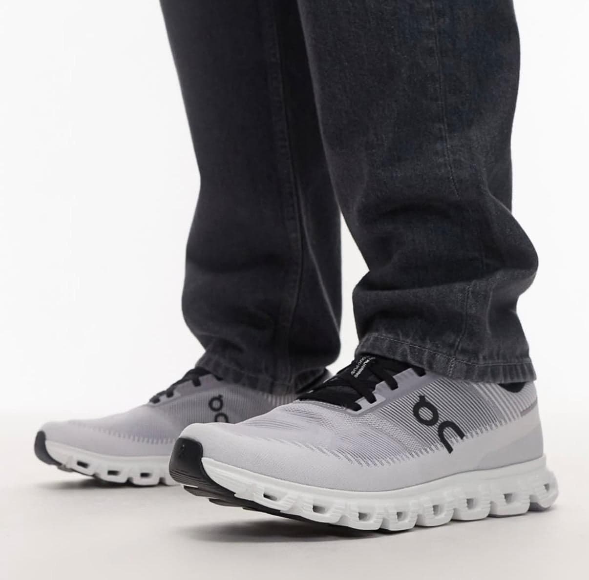 d282fb43-4efc-47e4-8ca5-ea3c60a5cff3 | AE JERSEY Light-gray and white running shoes with black laces worn with dark jeans, standing on a light surface.