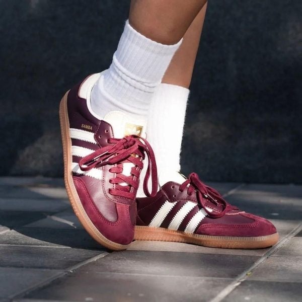 Maroon Adidas Samba sneakers with white stripes and laces, worn with white socks, standing on pavement.