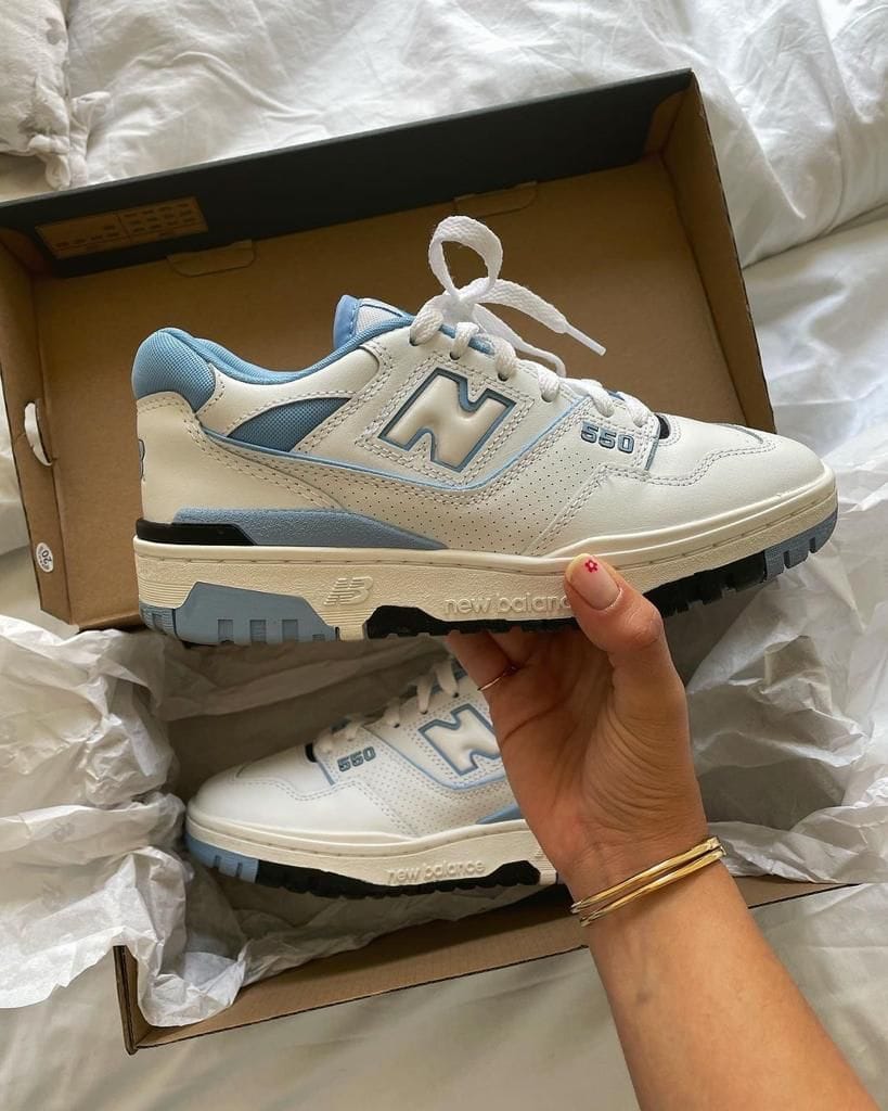 d471aca0-44db-4de2-857a-4a07d7dc0b4d_5dab3304-65bc-4b09-b44f-dc1ee0f5c523 | AE JERSEY White and light blue New Balance 550 sneaker being held over its shoe box, showing the side with the large 'N' logo and perforated panels.