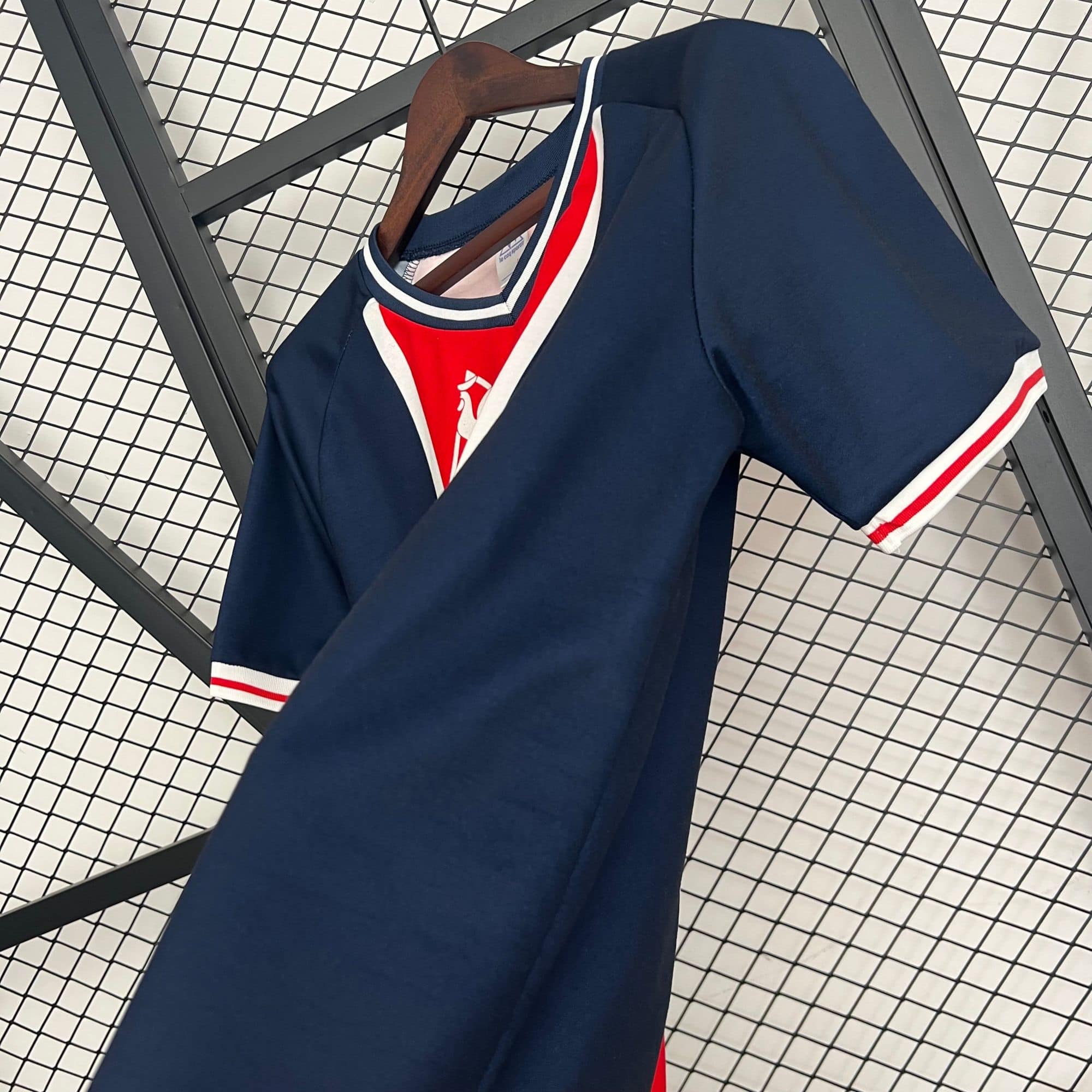 d4bc1a55_e114bb01-b960-498c-b85f-ae7a7ad8acfb | AE JERSEY Navy blue varsity-style jacket on a wooden hanger, with white and red trim, displayed against a metal grid backdrop in a store setting.