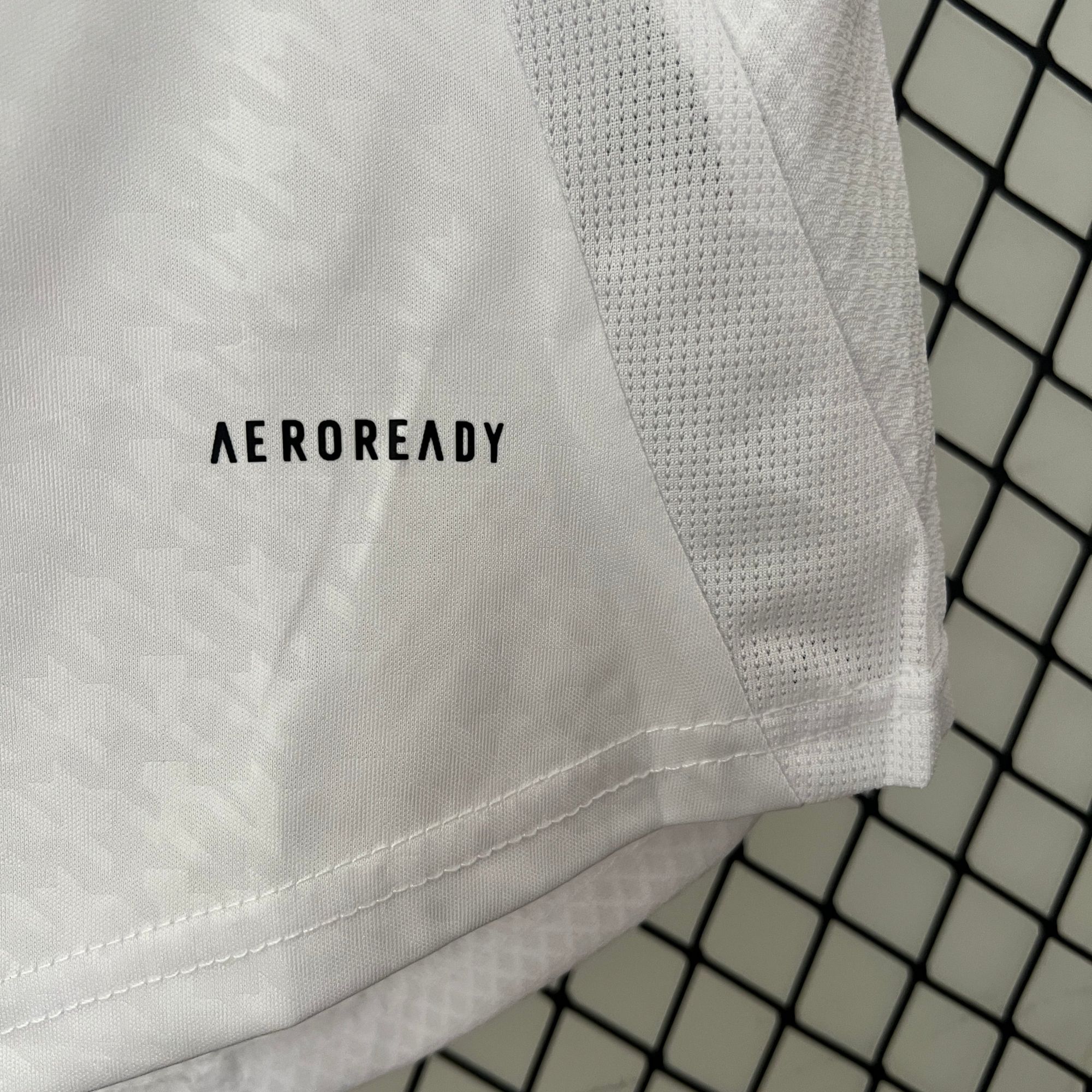 dfbba04e | AE JERSEY Close-up of white fabric with stitched seams and the word AEROREADY visible, set against a dark, tiled background grid.